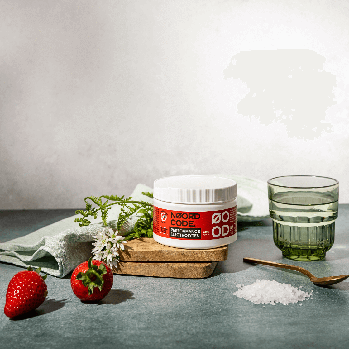 Červeno biela pixla elektrolytov na prkénku, pod nimi lyžička a vľavo jahody a kvetina s drobnými kvetmi, všetko na neutrálnom pozadí.